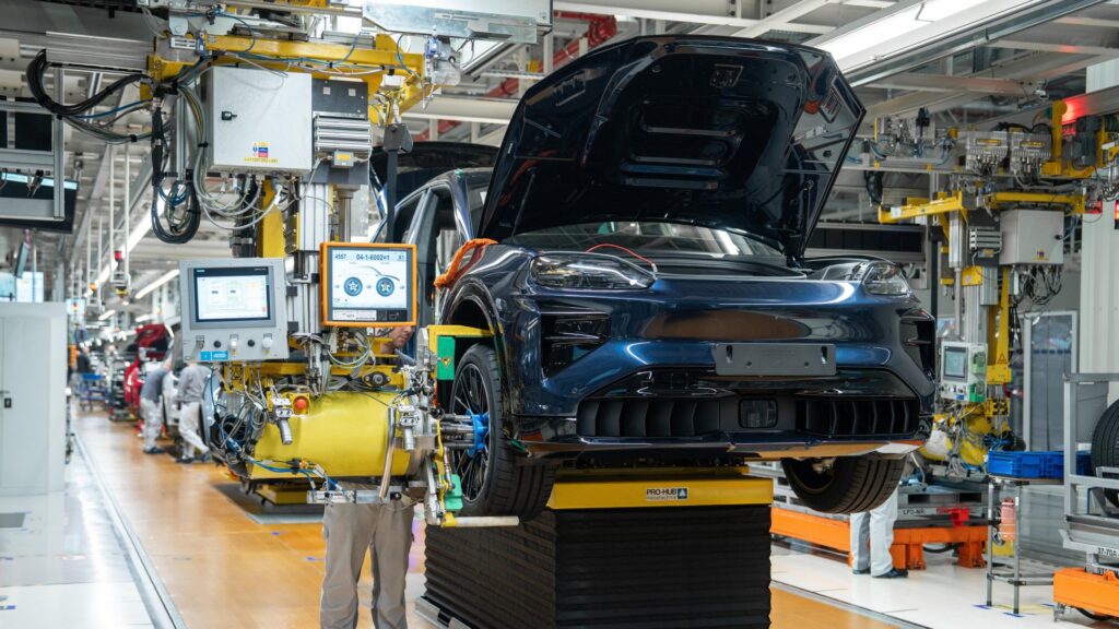 Um SUV Porsche Cayenne elétrico sendo montado na linha de produção da fábrica, com maquinário robótico ao redor, ilustrando a integração moderna de um powertrain elétrico no chassi do veículo.